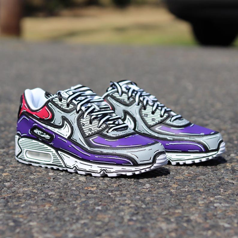Puede incluir: Un par de zapatillas moradas, grises y blancas con contornos negros. Los zapatos tienen un dise&ntilde;o &uacute;nico con un acento rojo en el tal&oacute;n. Las zapatillas est&aacute;n sobre una superficie de asfalto.