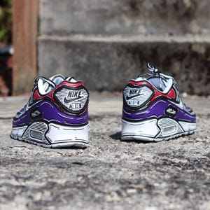 Puede incluir: Un par de zapatillas Nike Air Max 90 moradas, blancas y rojas con detalles de contorno negro. Las zapatillas presentan el logotipo de Nike en el tal&oacute;n y una suela blanca. Las zapatillas est&aacute;n sobre una superficie gris texturizada.