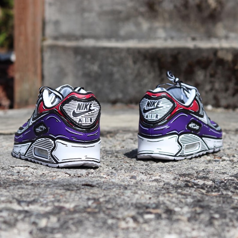 Puede incluir: Un par de zapatillas Nike Air Max 90 moradas, blancas y rojas con detalles de contorno negro. Las zapatillas presentan el logotipo de Nike en el tal&oacute;n y una suela blanca. Las zapatillas est&aacute;n sobre una superficie gris texturizada.