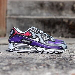 Puede incluir: Un par de zapatillas Air Max 90 con un dise&ntilde;o de c&oacute;mic. El zapato presenta una combinaci&oacute;n de colores p&uacute;rpura, gris, rojo y blanco con contornos negros. El texto "AIR MAX" es visible en el lateral.
