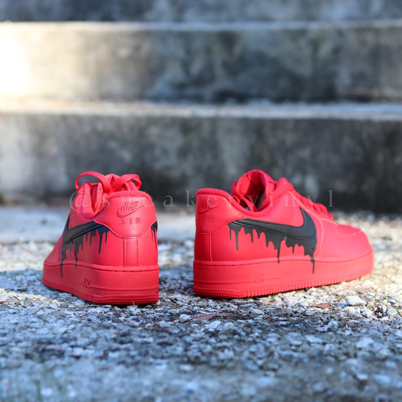 May include: Pair of red Nike Air Force 1 sneakers with black dripping paint design. The sneakers are on a stone surface with a concrete background. The Nike logo is visible on the side and heel.
