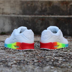 May include: White Nike Air Max 90 trainers with a rainbow-coloured outsole and red accents. The shoes feature the Nike Air logo on the side and the word "Air" on the back.
