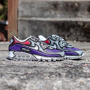 Puede incluir: Par de zapatillas Air Max 90 con un estilo de dibujos animados &uacute;nico. Los zapatos presentan una combinaci&oacute;n de colores p&uacute;rpura, gris, rojo y blanco con contornos negros. El dise&ntilde;o incluye el logotipo "Air Max" en el lateral.