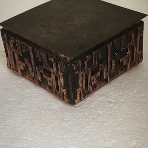 Könnte beinhalten: Eine quadratische Schachtel mit einem flachen, dunkelbraunen Deckel und Seiten, die ein erhabenes, strukturiertes, kupferfarbenes geometrisches Muster aufweisen. Die Schachtel hat eine metallische Oberfläche mit tiefen Vertiefungen zwischen den Elementen.