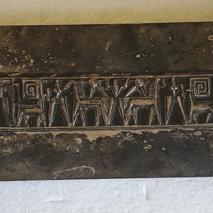 May include: A rectangular dark metal box with a weathered, mottled finish. The lid features a central horizontal relief panel depicting a series of stylized figures, animals, and geometric spiral patterns in a raised, metallic silver-toned design.