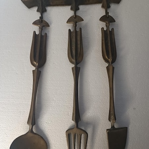 May include: A set of three antique-style brass kitchen utensils hanging on a wall-mounted rack. The utensils include a spoon, fork, and knife.