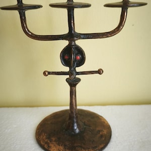 Puede incluir: Una escultura de metal en tono bronce de una figura estilizada con tres candelabros. La figura tiene una base redonda, un tallo central y una barra horizontal con dos pequeñas esferas. La cara de la figura tiene detalles rojos.
