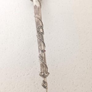 Może przedstawiać: Srebrny i biały ozdobny wskaźnik z wzorem Gwiazdy Dawida. Wskaźnik ma końcówkę w kształcie dłoni i dwie ozdobne kule na górze.