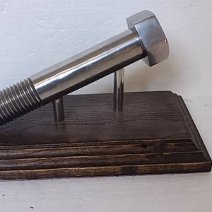 May include: A large, shiny metal bolt, mounted on a dark stained wooden base. The bolt is supported by two metal rods, creating a unique display piece. The bolt's threads and hexagonal head are clearly visible.