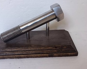Könnte beinhalten: Ein großer, glänzender Metallbolzen, montiert auf einem dunkel gebeizten Holzsockel. Der Bolzen wird von zwei Metallstangen getragen, wodurch ein einzigartiges Ausstellungsstück entsteht. Die Gewinde und der Sechskantkopf des Bolzens sind deutlich sichtbar.