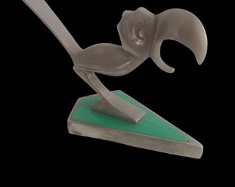 Op de afbeelding: Een gestileerde metalen sculptuur van een vogel met een grote snavel, geplaatst op een groene driehoekige basis. De vogel is afgebeeld in een minimalistische stijl, met een strakke, zilver-grijze afwerking. De sculptuur staat tegen een zwarte achtergrond.