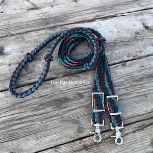 May include: Braided horse reins in navy blue, teal, and rust colors. The reins are coiled on a weathered wooden surface. The ends feature silver-colored metal clips. The text "Two Bay Ranch Co." is visible.