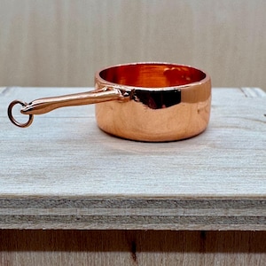 May include: A small, copper-coloured saucepan with a long handle and a ring at the end. The pot is round with a shiny finish, reflecting the light. The handle is attached to the side of the pot. The saucepan is sitting on a wooden surface.