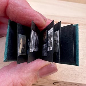 May include: Miniature open book with black pages and small black and white images. The cover is dark green. The book is held in a person's hand. The background is a light wood surface.