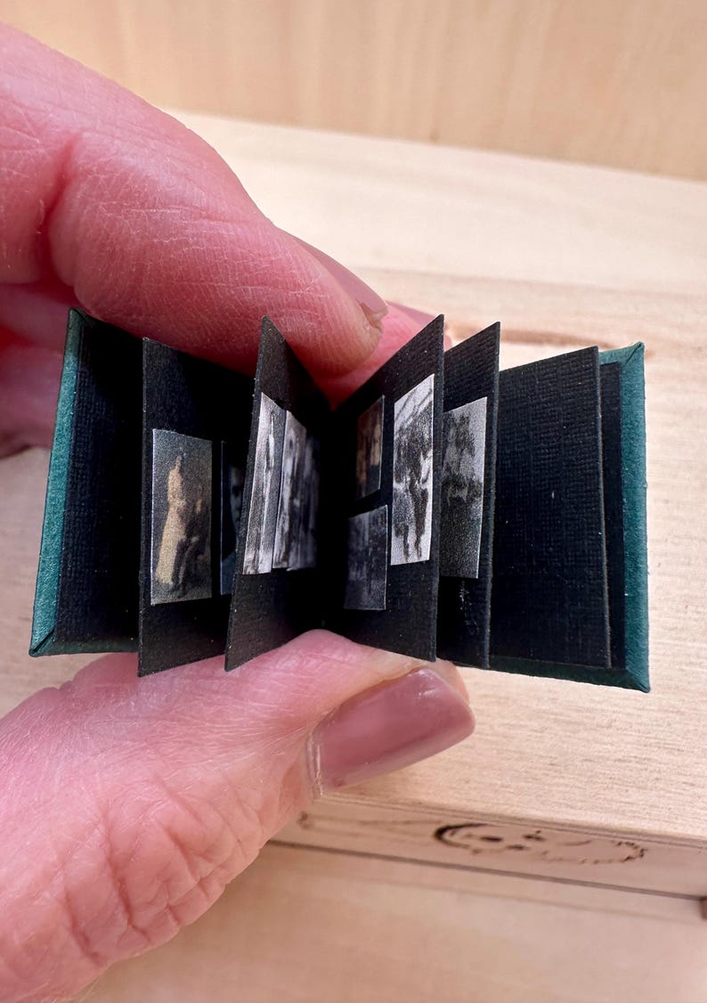 May include: Miniature open book with black pages and small black and white images. The cover is dark green. The book is held in a person's hand. The background is a light wood surface.