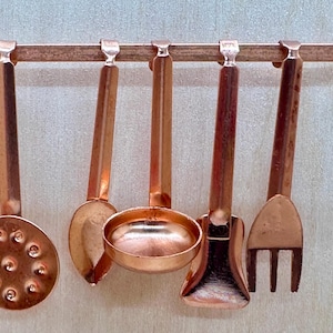 May include: A set of five copper kitchen utensils hanging on a metal rack. The utensils include a slotted spoon, a ladle, a small saucepan, a spatula, and a fork.
