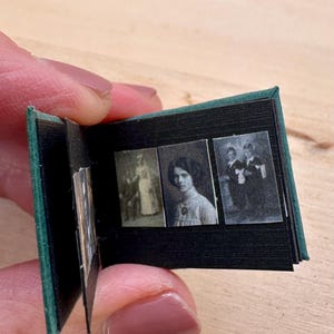 May include: Miniature photo album with a dark green cover and black pages. The album contains three small black and white photographs of people. The album is held open, displaying the images.