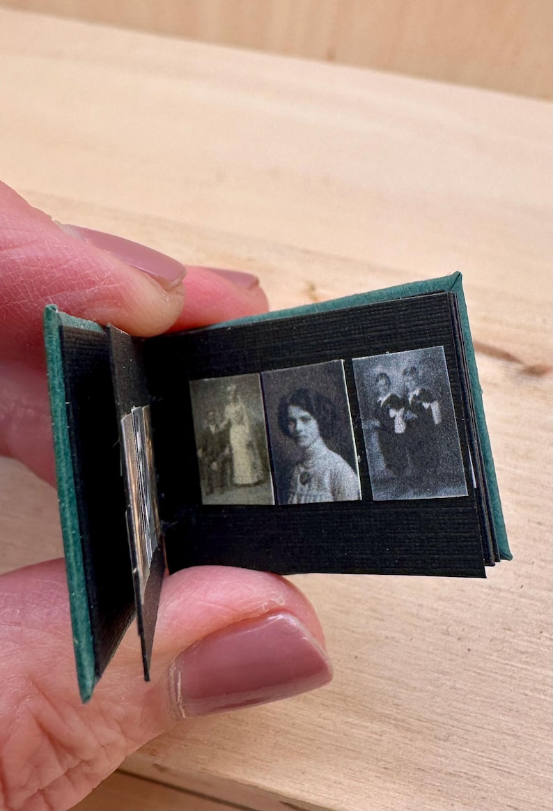 May include: Miniature photo album with a dark green cover and black pages. The album contains three small black and white photographs of people. The album is held open, displaying the images.