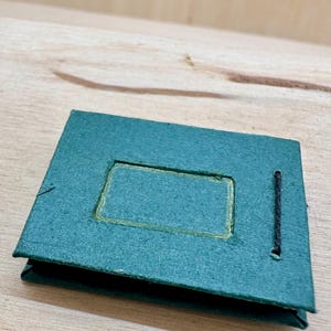 May include: A small, teal-colored photo album with a rectangular cutout on the front. The cutout is outlined in gold. A black string is attached to the side. The album is sitting on a light-colored wooden surface.
