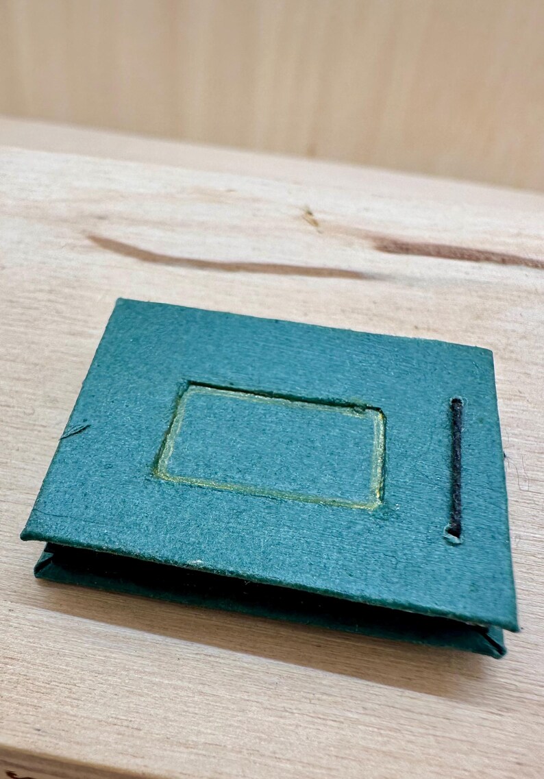 May include: A small, teal-colored photo album with a rectangular cutout on the front. The cutout is outlined in gold. A black string is attached to the side. The album is sitting on a light-colored wooden surface.