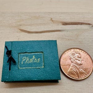 May include: A miniature teal photo album with the word "Photos" in gold lettering. A small black figurine is attached to the album. A penny is next to the album for size comparison.