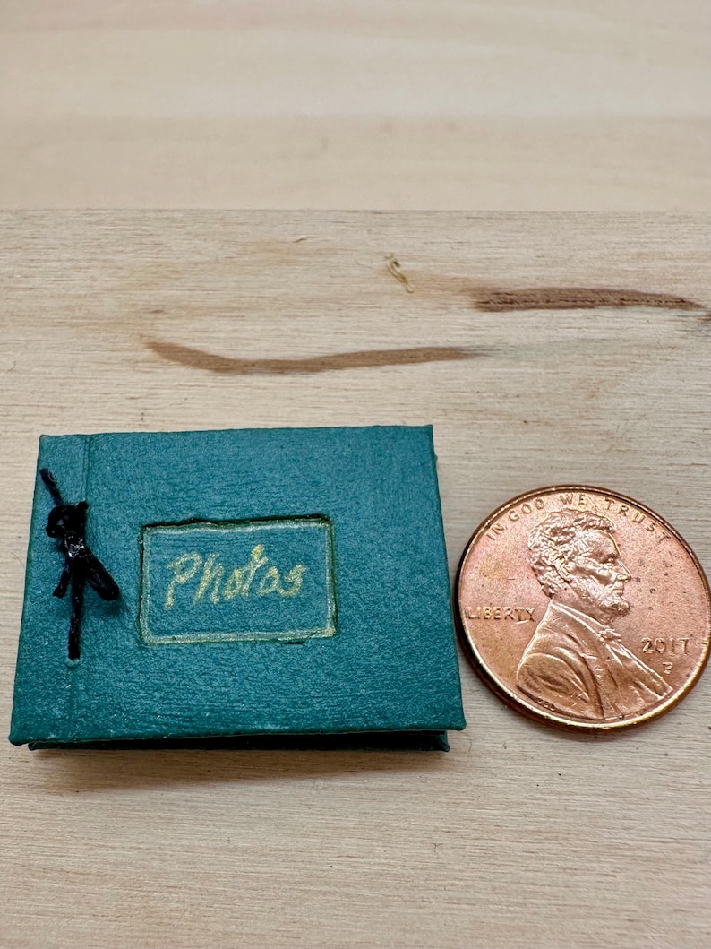 May include: A miniature teal photo album with the word "Photos" in gold lettering. A small black figurine is attached to the album. A penny is next to the album for size comparison.