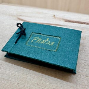 May include: A small, teal photo album with the word "Photos" in gold lettering. The album is rectangular and bound with a black string. The album is on a wooden surface.