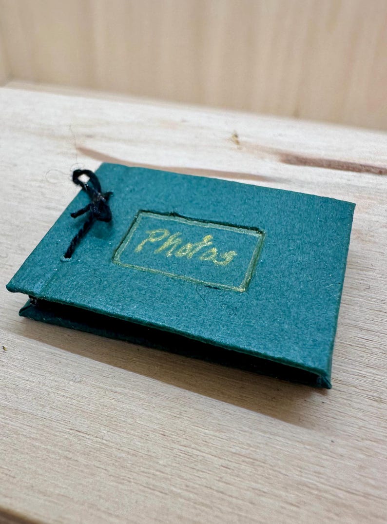 May include: A small, teal photo album with the word "Photos" in gold lettering. The album is rectangular and bound with a black string. The album is on a wooden surface.