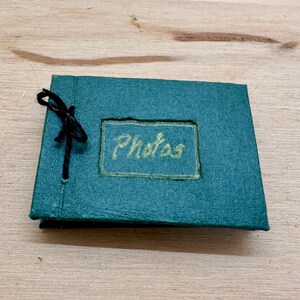 May include: A small, teal photo album with the word "Photos" in gold lettering. The album is tied with a black string. The album is on a light brown wooden surface.