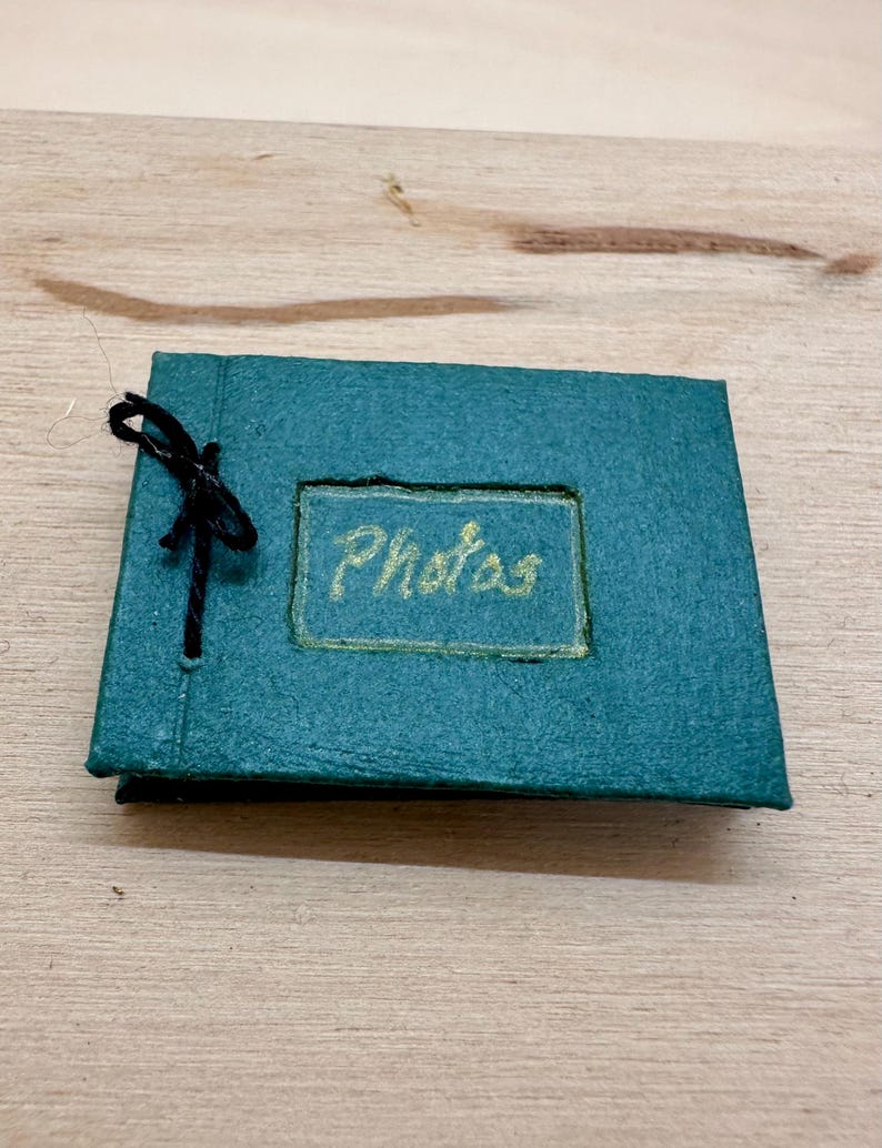 May include: A small, teal photo album with the word "Photos" in gold lettering. The album is tied with a black string. The album is on a light brown wooden surface.