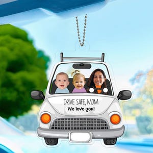 May include: A clear acrylic car ornament featuring a white car with three people inside. The text "DRIVE SAFE, MOM We love you!" is printed on the car. The ornament hangs from a silver chain.
