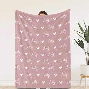 May include: A dusty rose-colored blanket featuring a repeating pattern of the name Lily written in a white cursive script, interspersed with small white heart icons arranged in a grid-like layout across the entire surface of the fabric.