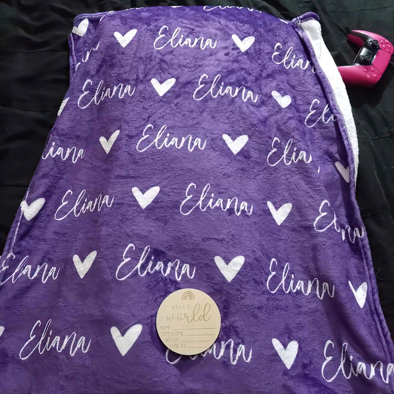 以下が含まれることがあります： 白のハートと「Eliana」という名前が全体に繰り返された紫色のフリースブランケット。右下には「Hello World」と書かれた木製の看板があります。