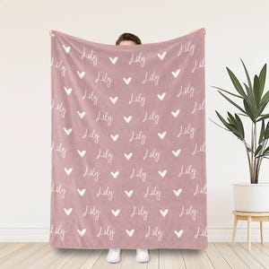 Op de afbeelding: Een oudroze deken met een herhalend patroon van de naam Lily in een wit cursief lettertype, afgewisseld met kleine witte hartjes die in een rasterachtig patroon over het gehele oppervlak van de stof zijn verdeeld.