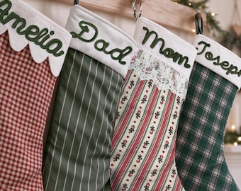 May include: Four Christmas stockings hanging in a row. The first is red and white gingham with the name Amelia. The second is olive green with white pinstripes and the name Dad. The third has candy cane stripes and lace with the name Mom. The fourth is green plaid with the name Joseph.