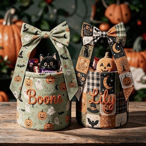 May include: Two cylindrical fabric Halloween baskets with large bows. The left basket is sage green with ghosts, pumpkins, and bats, featuring the name Boone. The right basket has a patchwork pattern of pumpkins, cats, and moons, featuring the name Lily.