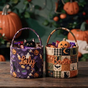 May include: Two cylindrical fabric Halloween baskets with handles. The left basket is purple with a pattern of pumpkins, ghosts, and cats, featuring the name Lily. The right basket has a patchwork design with Halloween motifs and the name Boone.