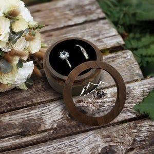 May include: A round wooden ring box with a clear lid, showcasing two rings on a black velvet lining. The lid has a mountain design and the names "Amelia + Liam" with a date. The box is on a weathered wooden surface, with a floral bouquet in the background.
