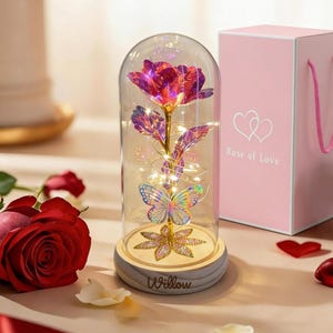 May include: A decorative glass dome with a metallic pink rose, butterfly, and fairy lights. The base is wooden with the word "Willow" engraved. A pink gift box with the text "Rose of Love" is next to the dome.