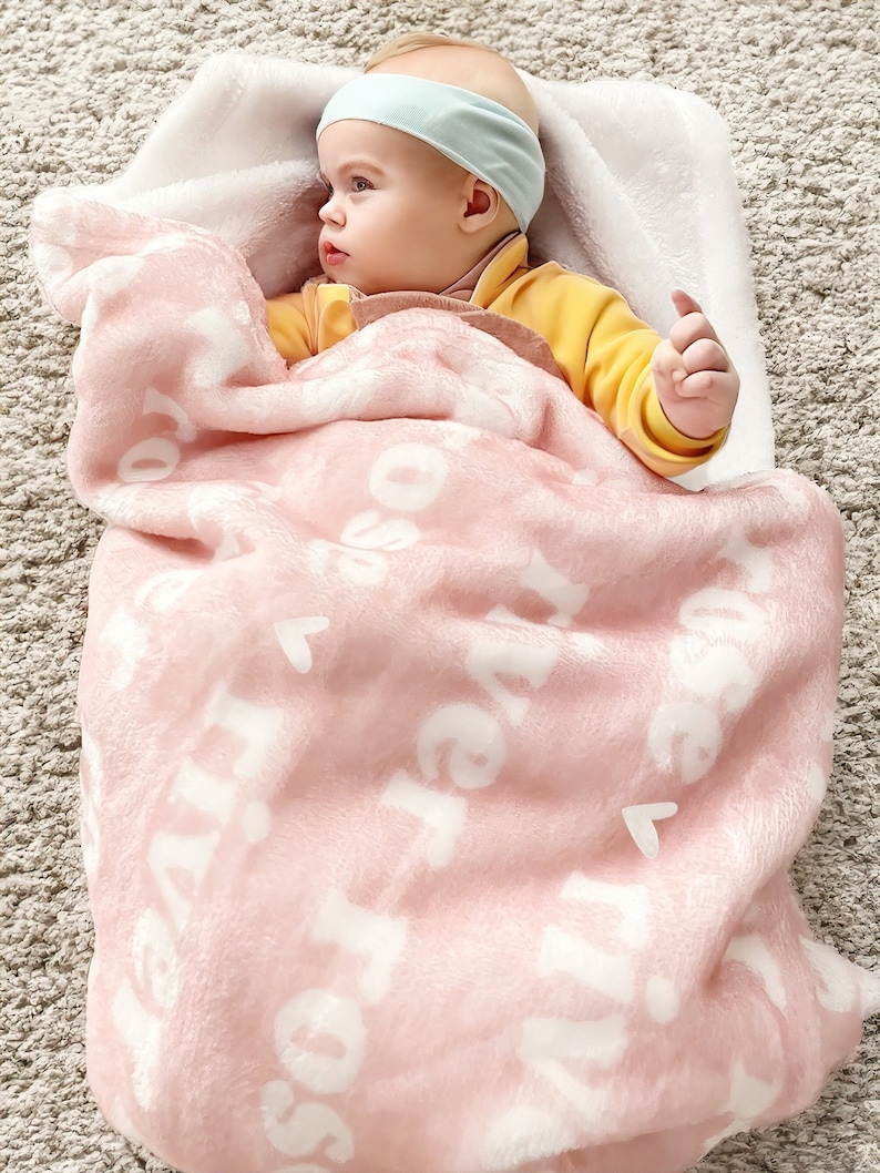 May include: A pink baby blanket with white letters that spell out "Love" repeatedly. A baby is lying on the blanket, wearing a yellow shirt and a light blue headband.