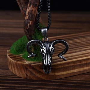 Könnte beinhalten: Halskette mit einem silberfarbenen Widder-Schädel-Anhänger. Der Anhänger zeigt ein detailliertes Widder-Schädel-Design mit gebogenen Hörnern. Die Halskette hat eine dunkle Kette und wird auf einer Holzoberfläche mit Moos präsentiert.