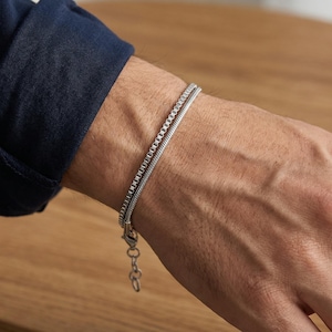 Könnte beinhalten: Ein silbernes Armband mit zwei verschiedenen Kettenstilen. Eine Kette hat ein quadratisches Glieddesign, die andere ein abgerundetes, schlangenartiges Aussehen. Das Armband befindet sich an einem Handgelenk, mit einem marineblauen Hemdärmel im Hintergrund.