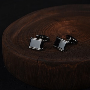 Puede incluir: Un par de gemelos rectangulares negros con un acabado brillante y un borde de pequeños detalles oscuros. Los gemelos se exhiben sobre una superficie de madera marrón oscuro, creando un contraste.