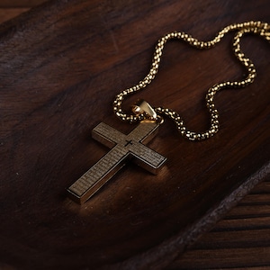 以下が含まれることがあります： 詳細なデザインのゴールドカラーのクロス ペンダント ネックレス。クロスは長方形で、中央に小さなクロスがエッチングされています。ネックレスチェーンは同じゴールドカラーで、濃い木製の表面に置かれています。