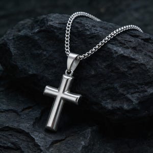 Puede incluir: Un collar con colgante de cruz plateada. La cruz tiene un diseño simple y clásico con bordes redondeados, suspendida de una cadena a juego. El collar se exhibe sobre una superficie oscura y texturizada.
