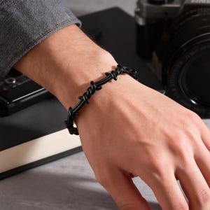 May include: A black barbed wire bracelet is displayed on a wrist. The bracelet is made of linked, curved segments, creating a spiky appearance. The jewelry is set against a neutral background, with a hint of a camera and a dark-colored book.