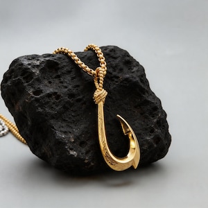 Puede incluir: Un colgante de anzuelo de metal en tono dorado suspendido de una cadena de eslabones cuadrados a juego. El colgante presenta un intrincado patrón floral grabado a lo largo de su superficie curva y está unido a la cadena con un detalle de bucle de metal anudado.