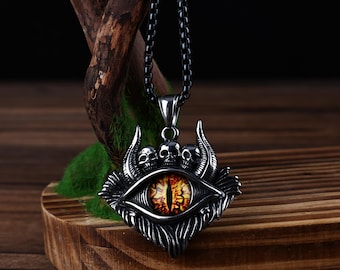 May include: A silver-colored pendant necklace featuring a detailed eye design with a yellow and orange iris. The pendant includes three small skulls and decorative elements. The necklace has a black chain and is displayed on a wooden stand.