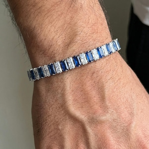 May include: A silver bracelet with alternating rectangular blue and clear gemstones. The bracelet is worn on a wrist, showcasing the jewelry's design and color contrast. The gemstones are set closely together, creating a continuous pattern.