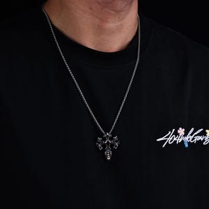 May include: Silver-toned cross pendant necklace. The pendant features an ornate design with intricate details. The necklace is worn on a black t-shirt. The shirt has the text "404 Not Found Gang" in a cursive font.